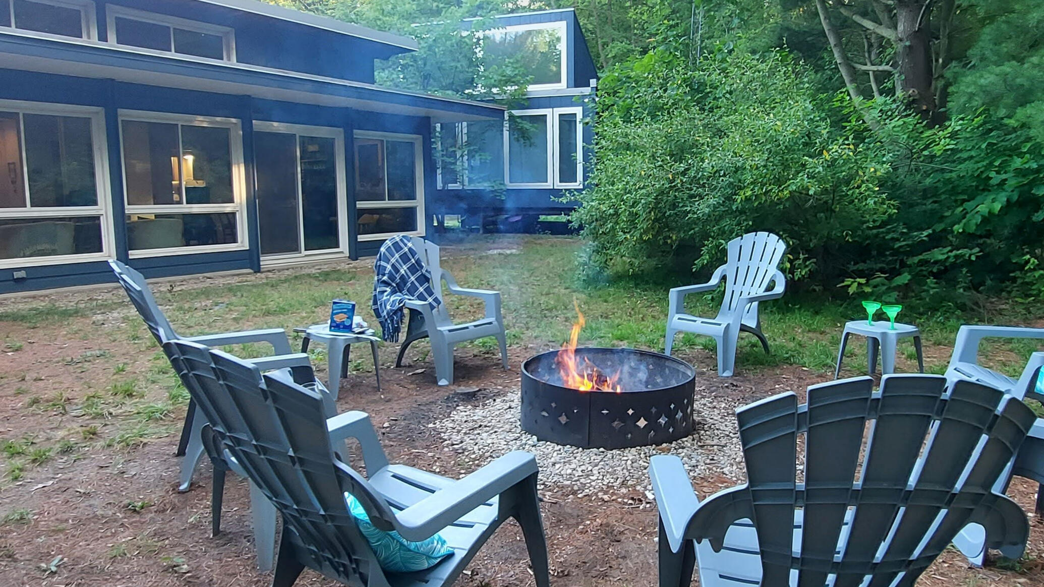 chairs around firepit with fire going, wooded area and blue house in background